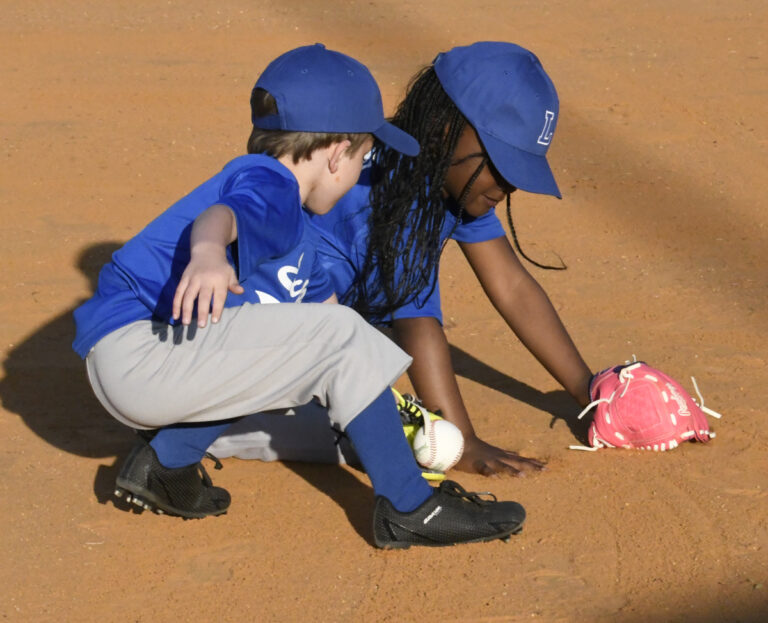 Two young children in blue baseball uniforms and hats crouch on a baseball field, reaching for a ball on the ground with their gloves.