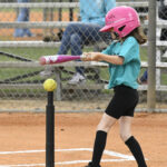A young girl wearing a pink helmet swings at a baseball on a tee during a tee-ball game, with a fence and spectators in the background.