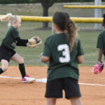 Young girls in green shirts playing softball on a dirt field, one catching a ball while others watch and get ready to play.