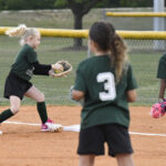 Young girls in green uniforms playing softball on a field, one preparing to catch a ball while others watch and participate in the game.