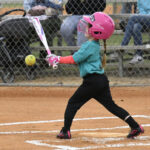 A young girl in a pink helmet hits a softball with a bat on a baseball field, with a fence and spectators in the background.