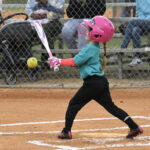 A young child in a pink helmet swings a bat at a softball on a baseball field, with spectators behind a fence.