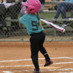Young girl wearing a pink helmet and turquoise jersey swings a baseball bat during a game, standing on a dirt field with a chain-link fence in the background.
