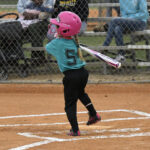 A young girl wearing a pink helmet and teal jersey swings a bat during a softball game, with spectators sitting on bleachers behind a chain-link fence.