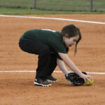 A young softball player in a green shirt crouches on a dirt field, fielding a ground ball with a glove.
