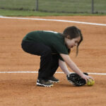 A young girl in a green shirt and black pants fielding a softball on a dirt infield during a game.