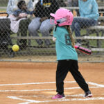 Young girl in a teal shirt and pink helmet swings a bat at a softball during a game, with spectators watching from behind a fence.