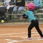Young child in a teal shirt and pink helmet swinging a bat at a pitched softball on a baseball field, with spectators in the background.