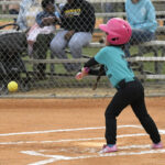 A young child wearing a pink helmet swings a bat at a softball during a game, with spectators watching from behind a fence in the background.