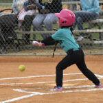A young child in a pink helmet attempts to hit a pitched softball on a field, while spectators watch from the stands behind a chain-link fence.