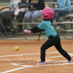 A young girl wearing a pink helmet swings a bat at a softball on a baseball field, with spectators watching from behind a chain-link fence.