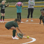 Young girls playing softball on a dirt field, one bending down to catch a ground ball while others watch and prepare in the background.