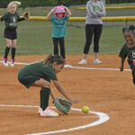 Young girls play softball on a field; one bends down to catch a rolling ball while others watch or prepare to run.
