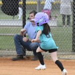 A young girl wearing a purple helmet runs on a softball field, while an adult sits near the fence in the background.