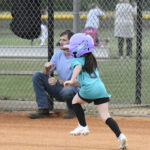 A young girl wearing a purple helmet runs on a baseball field while a man sits nearby, with a fence and people visible in the background.