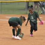 Two young girls in green shirts and black shorts play softball on a dirt field, one bending to pick up a ball while the other smiles nearby.