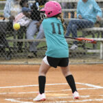 A young girl wearing a pink helmet and teal shirt prepares to hit a softball during a game, with spectators in the background.