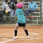A young girl wearing a pink helmet and teal shirt prepares to bat at a softball game, with spectators watching behind a chain-link fence.
