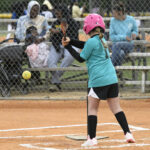 A young girl in a teal shirt and pink helmet prepares to bat at a softball game, with spectators watching from bleachers behind a fence.