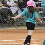 A young girl wearing a teal shirt, black shorts, and a pink helmet runs on a baseball field, with spectators visible in the background behind a chain-link fence.