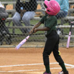 Young child in a pink helmet swings a bat during a youth baseball game, with spectators sitting on bleachers in the background.