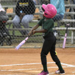 A young child wearing a pink helmet swings a bat during a baseball game, with spectators sitting on benches in the background.