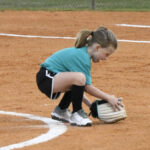 Young girl in sportswear crouches on a baseball field, fielding a ground ball with a glove near a white line.