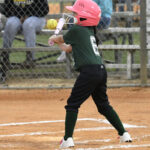 A young child wearing a pink helmet and green uniform swings a bat at a softball during a game, with spectators seated in the background.