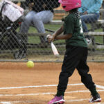 A child in a green shirt and pink helmet swings at a softball on a dirt field, with spectators sitting behind a chain-link fence.