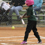 A child in a pink helmet prepares to bat at a baseball game, facing an incoming pitch, with spectators sitting on bleachers in the background.