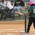 A young child wearing a pink helmet prepares to hit a baseball from a tee during a game, with spectators watching behind a chain-link fence.