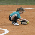 A young girl squats on a softball field, picking up a yellow softball with a glove, wearing a teal shirt, black shorts, and knee-high socks.