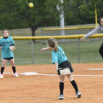 Young girls play softball on a dirt field, one pitching the ball while another stands ready. A coach or adult observes in the background near the fence.