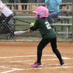 A young child wearing a pink helmet and green jersey prepares to swing a bat at a baseball game, standing at home plate on a dirt field.