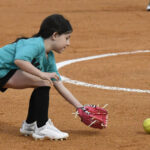 A young girl in a teal shirt and black shorts prepares to catch a softball with a red glove on a dirt field.