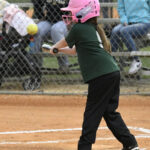 A young girl wearing a pink helmet and green shirt swings a bat at a softball during a game, with spectators in the background.