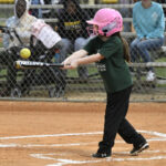 A young girl wearing a pink helmet hits a softball with a bat on a baseball field, spectators visible behind a chain-link fence.