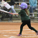A young girl wearing a purple helmet and green shirt swings a bat during a youth softball game, with spectators watching from behind a fence.