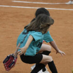Two young girls in teal shirts and black shorts playing softball, crouching on a dirt field, one holding a glove, preparing to field the ball.