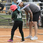 A coach gives batting tips to a young girl in a helmet during a little league baseball game.