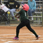 A young girl wearing a pink helmet swings at a softball during a game on a dirt field, with spectators watching behind a chain-link fence.