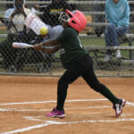 A young girl wearing a pink helmet hits a softball with a bat on a baseball field, while spectators watch from behind a chain-link fence.