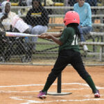 A child wearing a pink helmet swings a bat at a baseball set on a tee while people watch from the bleachers in the background.