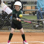 Young child in a helmet swings at a softball during a game, standing at bat on a dirt field with spectators in the background.