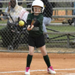 A young girl in a helmet prepares to hit a softball during a game, standing at bat with spectators and a fence in the background.