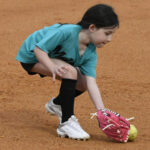 Young girl in teal shirt and black shorts fielding a softball with a pink glove on a dirt field.