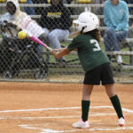 A young girl in a green jersey and white helmet swings a bat at a softball during a game, with spectators watching behind a fence.