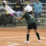 A young girl in a green jersey and white helmet bats at a softball game while spectators watch from bleachers behind a fence.