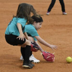 Two young girls in teal shirts field a ground ball on a baseball diamond, both reaching for the softball with their gloves.