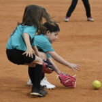 Two young girls in teal shirts playing softball, both reaching to catch a yellow softball on a dirt field.